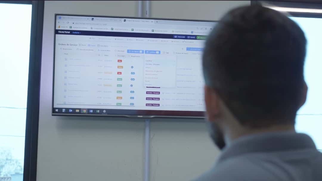 Homem visto de costas analisa os dados de ordens de serviço do CMMS TracOS™ em uma televisão fixada na parede.
